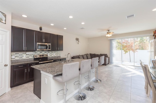 kitchen featuring sink, appliances with stainless steel finishes, a kitchen breakfast bar, light stone countertops, and a center island with sink