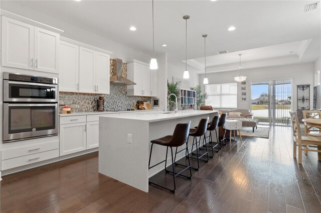 kitchen featuring white cabinets, a kitchen island with sink, wall chimney range hood, and a tray ceiling