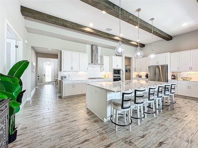 kitchen with a large island with sink, white cabinets, appliances with stainless steel finishes, wall chimney exhaust hood, and beamed ceiling