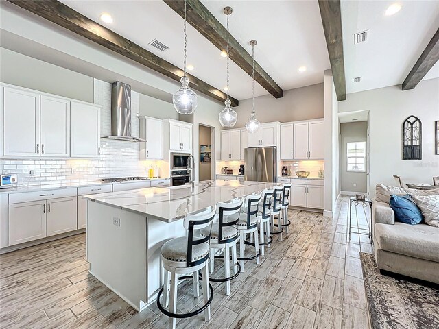 kitchen featuring appliances with stainless steel finishes, hanging light fixtures, wall chimney range hood, white cabinets, and beam ceiling