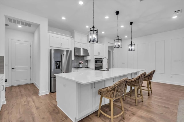 kitchen featuring a large island with sink, white cabinets, appliances with stainless steel finishes, hanging light fixtures, and a breakfast bar