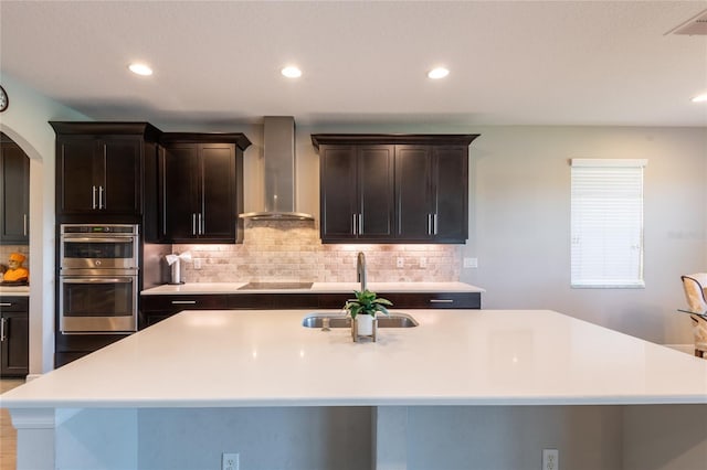 kitchen featuring a kitchen island with sink, sink, stainless steel double oven, and wall chimney range hood