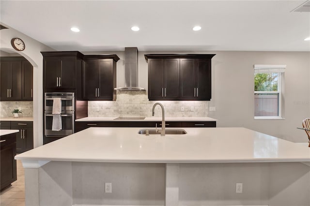 kitchen with an island with sink, wall chimney exhaust hood, double oven, and light countertops