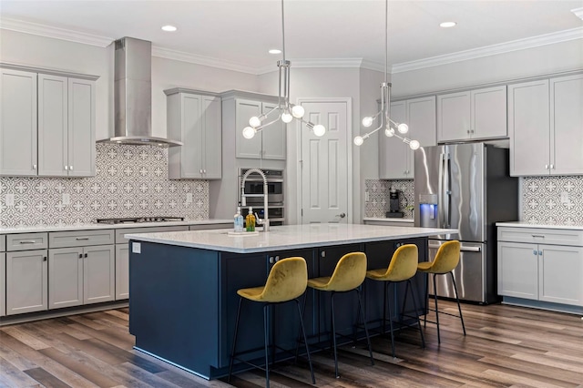 kitchen featuring wall chimney exhaust hood, backsplash, hanging light fixtures, a kitchen island with sink, and appliances with stainless steel finishes