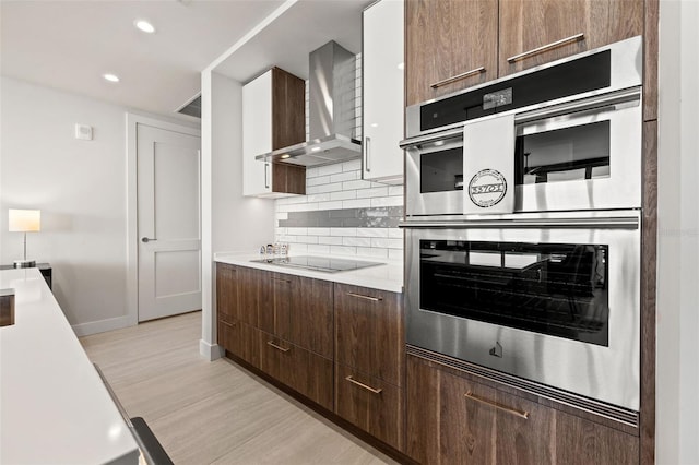 kitchen with stainless steel double oven, wall chimney exhaust hood, decorative backsplash, light hardwood / wood-style flooring, and black electric cooktop