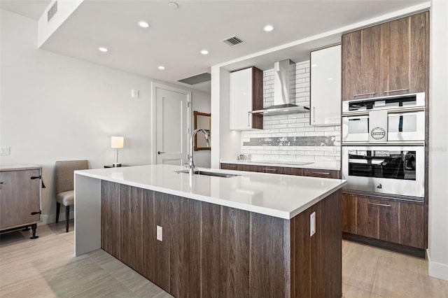 kitchen with an island with sink, sink, wall chimney range hood, and double oven