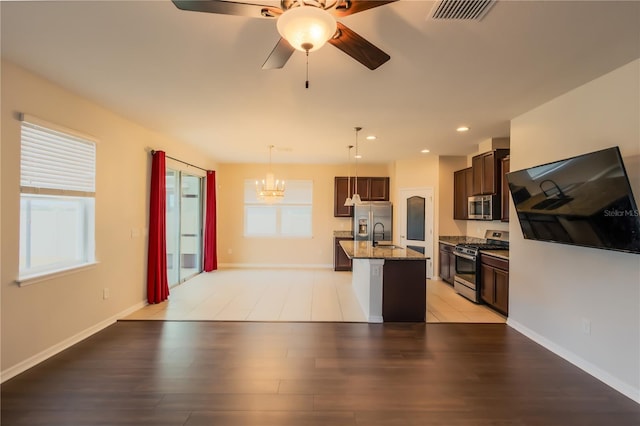 kitchen with an island with sink, appliances with stainless steel finishes, hanging light fixtures, light stone countertops, and ceiling fan with notable chandelier