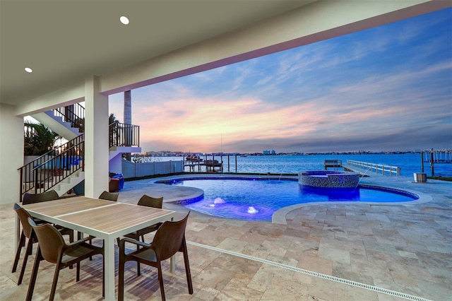 pool at dusk featuring an in ground hot tub, a water view, pool water feature, and a patio