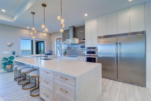 kitchen featuring an island with sink, sink, white cabinets, stainless steel appliances, and wall chimney exhaust hood