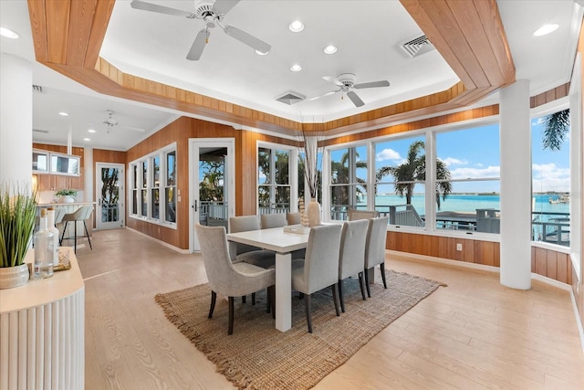 dining space with a water view, a tray ceiling, wood walls, and light hardwood / wood-style flooring