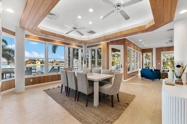 dining space featuring a water view, a tray ceiling, wooden walls, ceiling fan, and light hardwood / wood-style floors