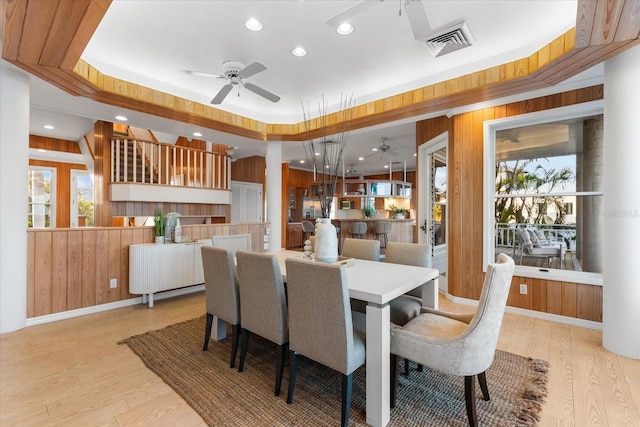 dining area with ceiling fan, wooden walls, a raised ceiling, and light hardwood / wood-style flooring