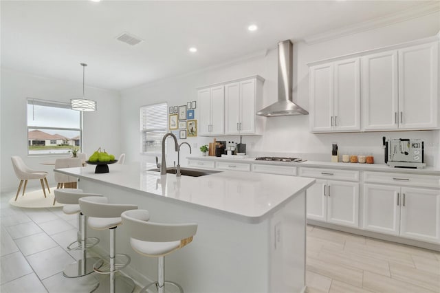 kitchen with hanging light fixtures, a kitchen island with sink, wall chimney range hood, and white cabinets