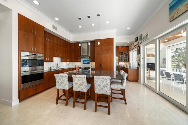 kitchen featuring a spacious island, wall chimney range hood, hanging light fixtures, and double oven