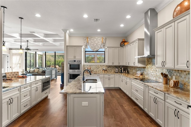 kitchen with coffered ceiling, an island with sink, appliances with stainless steel finishes, wall chimney range hood, and beam ceiling