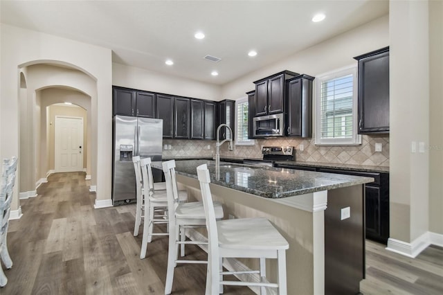 kitchen featuring an island with sink, dark stone counters, appliances with stainless steel finishes, and a kitchen bar