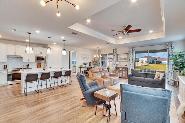 living room featuring ceiling fan with notable chandelier, light wood-type flooring, sink, and a tray ceiling