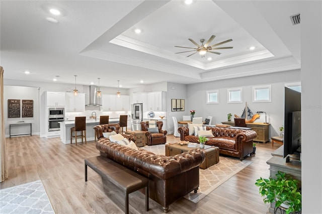 living room with ceiling fan, a raised ceiling, sink, and light wood-type flooring