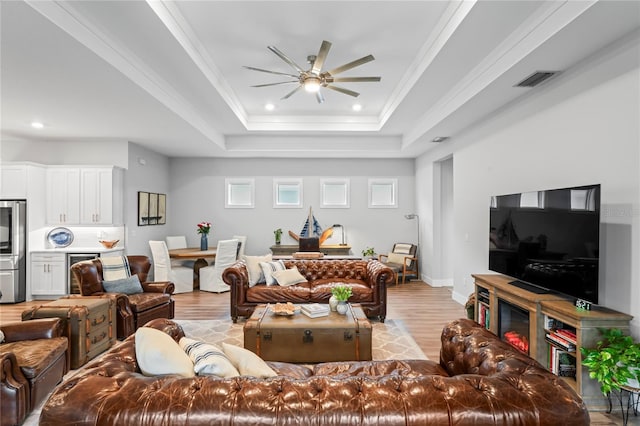 living room featuring ceiling fan, ornamental molding, a tray ceiling, and light wood-type flooring