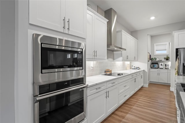 kitchen with white cabinets, stainless steel appliances, light hardwood / wood-style floors, decorative backsplash, and wall chimney range hood