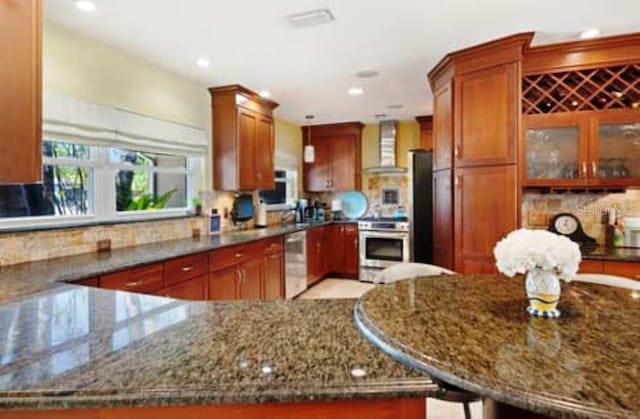 kitchen featuring appliances with stainless steel finishes, kitchen peninsula, stone counters, and wall chimney exhaust hood