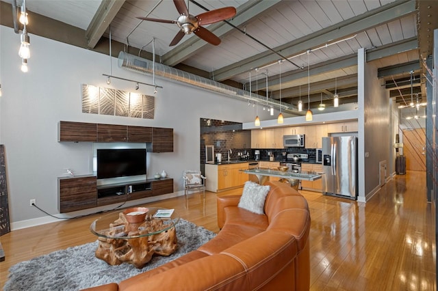 living room featuring ceiling fan, light wood-type flooring, beamed ceiling, and a high ceiling