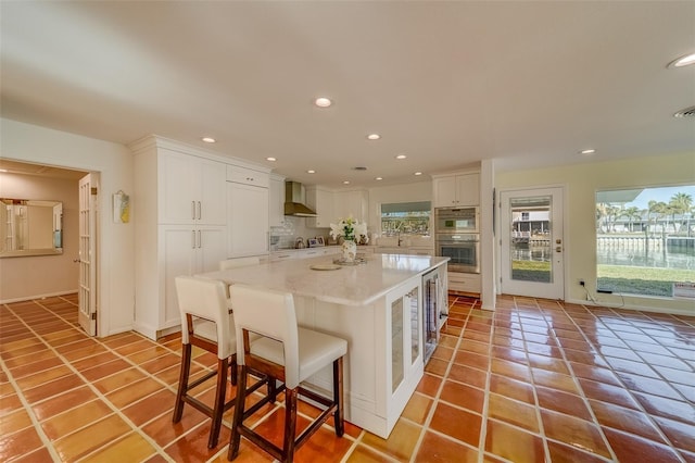 kitchen featuring wall chimney exhaust hood, a kitchen island, white cabinetry, stainless steel double oven, and a kitchen breakfast bar