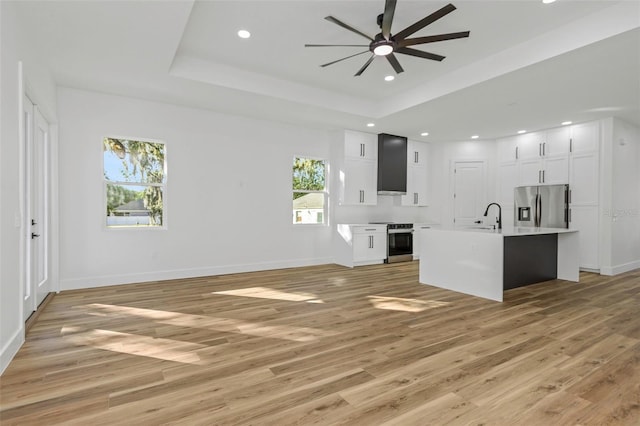 unfurnished living room with ceiling fan, sink, a tray ceiling, and light hardwood / wood-style flooring