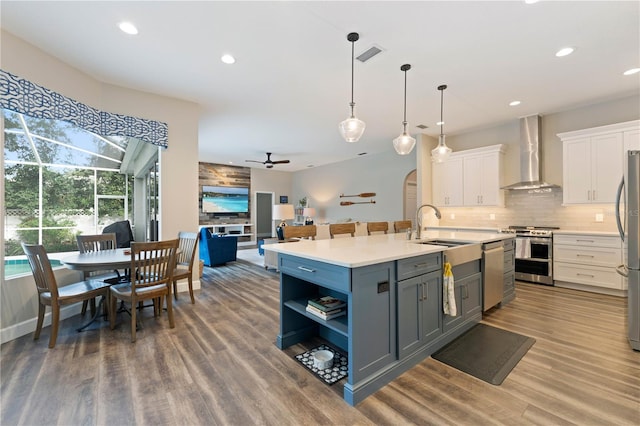 kitchen with a kitchen island with sink, stainless steel appliances, white cabinets, wall chimney range hood, and decorative light fixtures