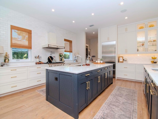 kitchen with white cabinetry, a center island with sink, built in refrigerator, decorative backsplash, and wall chimney exhaust hood