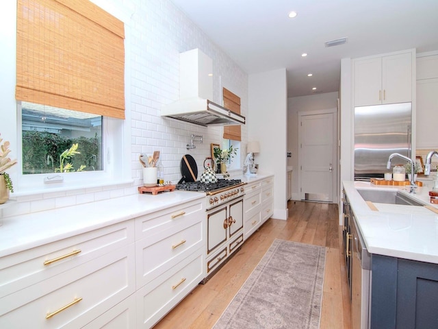 kitchen with built in fridge, white cabinets, wall chimney range hood, sink, and light hardwood / wood-style flooring