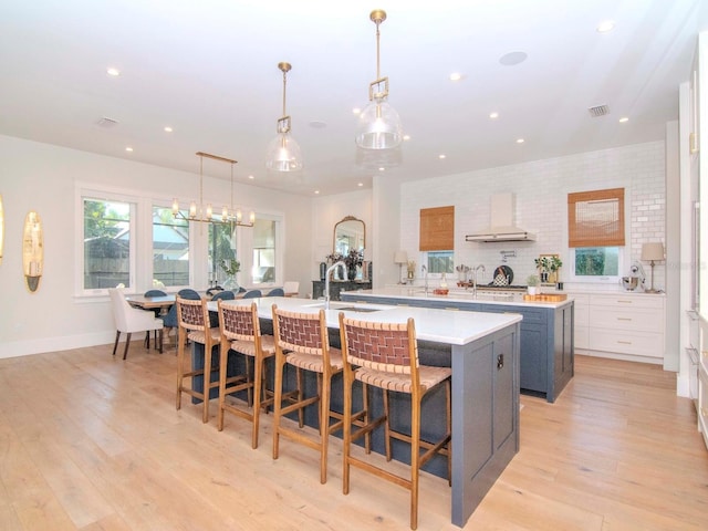 kitchen with white cabinets, a large island with sink, wall chimney range hood, and hanging light fixtures