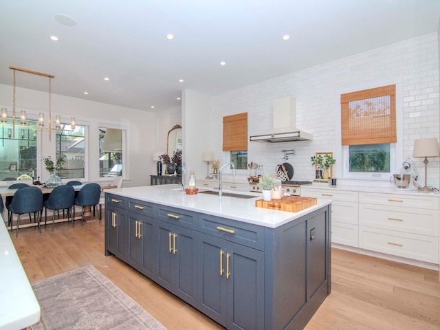 kitchen with wall chimney range hood, a center island with sink, sink, white cabinetry, and light wood-type flooring