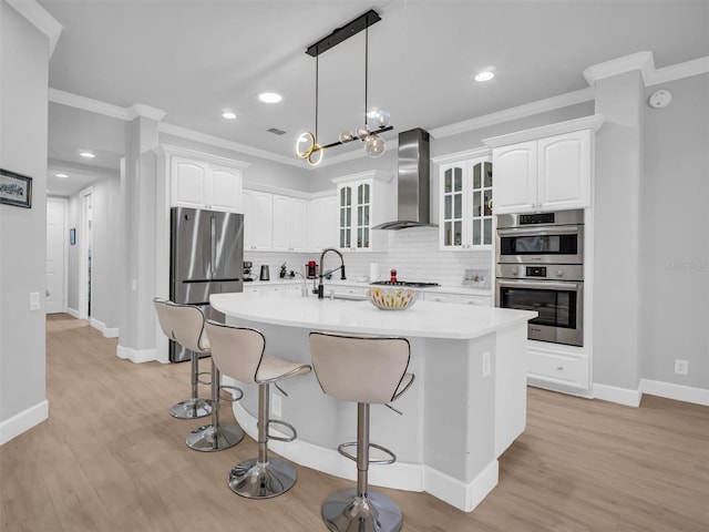 kitchen with wall chimney range hood, an island with sink, white cabinetry, and appliances with stainless steel finishes