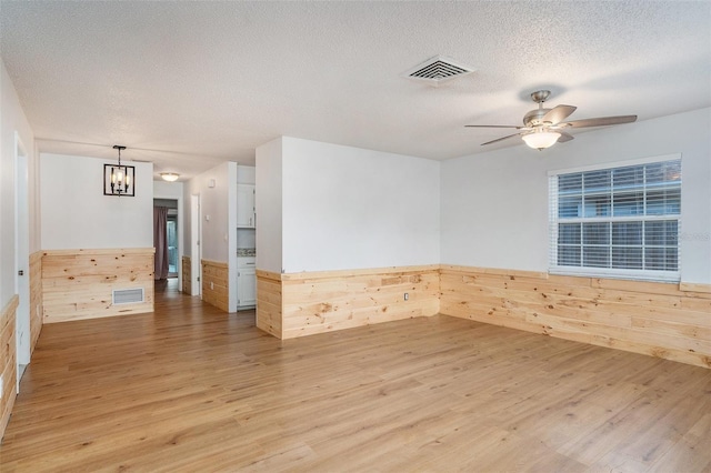 empty room with ceiling fan with notable chandelier, wooden walls, a textured ceiling, and light hardwood / wood-style flooring