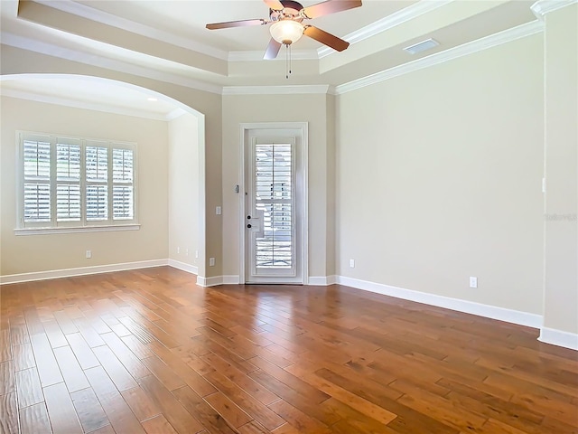 empty room featuring arched walkways, dark wood-style flooring, a raised ceiling, ornamental molding, and baseboards