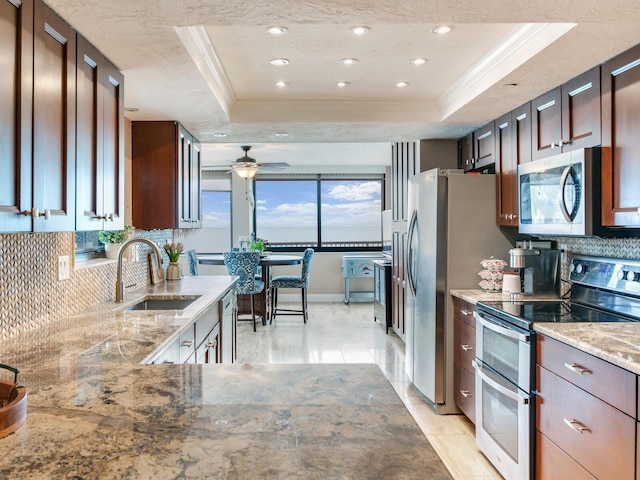 kitchen with ceiling fan, sink, a tray ceiling, light stone countertops, and stainless steel appliances