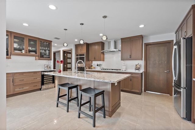 kitchen with wine cooler, pendant lighting, a kitchen island with sink, appliances with stainless steel finishes, and wall chimney exhaust hood