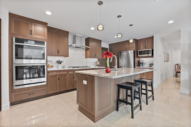 kitchen featuring light stone countertops, appliances with stainless steel finishes, wall chimney exhaust hood, hanging light fixtures, and a kitchen island with sink