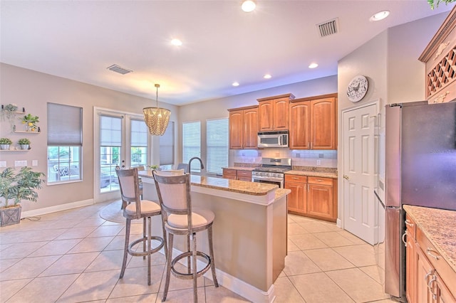 kitchen featuring hanging light fixtures, light tile patterned floors, an island with sink, stainless steel appliances, and light stone countertops