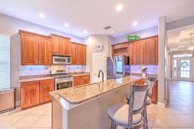 kitchen with sink, an island with sink, stainless steel appliances, light stone countertops, and decorative backsplash