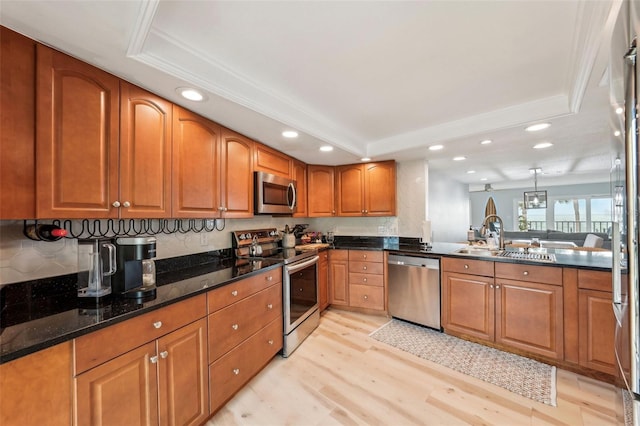 kitchen with appliances with stainless steel finishes, a raised ceiling, decorative light fixtures, dark stone countertops, and light hardwood / wood-style flooring