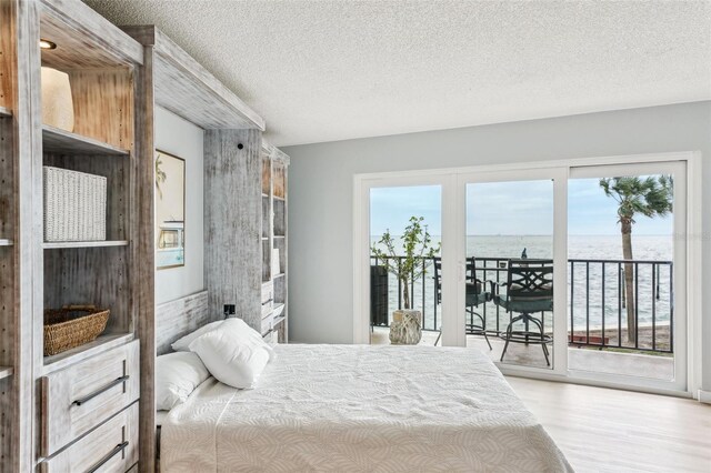 bedroom featuring a water view, access to exterior, light wood-type flooring, and a textured ceiling