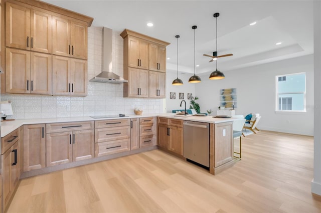 kitchen with wall chimney range hood, hanging light fixtures, a raised ceiling, stainless steel dishwasher, and kitchen peninsula