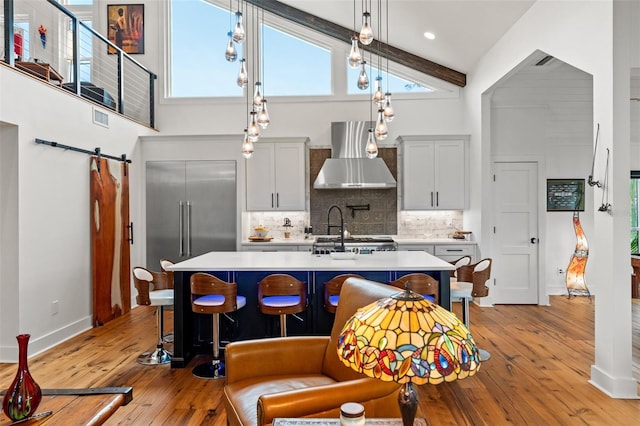 kitchen with a barn door, wall chimney range hood, beam ceiling, light hardwood / wood-style flooring, and a kitchen island with sink