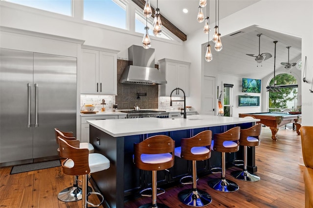 kitchen featuring built in refrigerator, wall chimney exhaust hood, pool table, an island with sink, and decorative backsplash