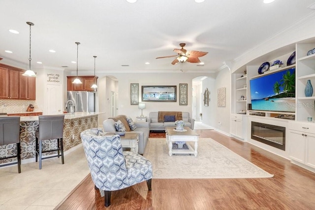 living room featuring ceiling fan, built in shelves, crown molding, light hardwood / wood-style flooring, and sink