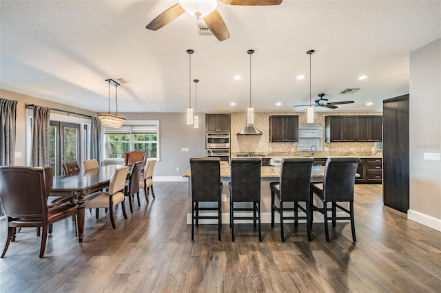 kitchen with wall chimney range hood, a kitchen island with sink, backsplash, a breakfast bar area, and dark brown cabinets