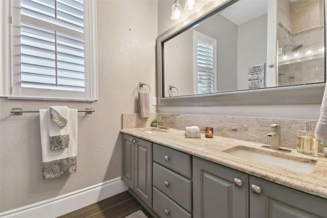 bathroom featuring wood finished floors, baseboards, double vanity, a tile shower, and a sink