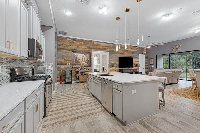 kitchen featuring stainless steel appliances, sink, wooden walls, a center island with sink, and pendant lighting